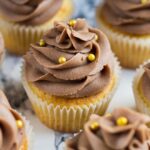 Chocolate buttercream frosted yellow cupcakes with gold ball sprinkles on marble background.