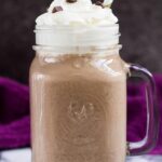 A mason jar filled with mocha smoothie topped with whipped cream and chocolate chips with straw.