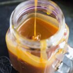 Overhead shot of spoon creating ripples in a mason jar full of salted caramel sauce on baking tray.