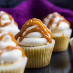 Frosted vanilla cupcakes topped with a drizzle of salted caramel sauce on a baking tray.