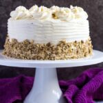 A cream cheese frosted carrot layer cake studded with crushed pecans on a white cake stand.