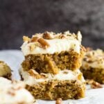A stack of two cream cheese frosted carrot cake blondies with a bite taken out of the top one on parchment paper.