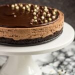 A no-bake chocolate cheesecake topped with chocolate ganache and chocolate chips on a white cake stand.