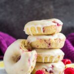A stack of baked lemon raspberry doughnuts with a bitten doughnut leaning up the side on parchment paper.