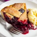 Slice of homemade cherry pie on a small white plate with a scoop of ice cream and a fork.
